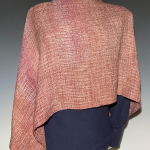 May include: A textured, woven shawl in a muted dusty rose color with a visible herringbone pattern. The shawl is draped over a dark navy blue long-sleeved shirt and black trousers, displayed on a mannequin torso against a neutral background.