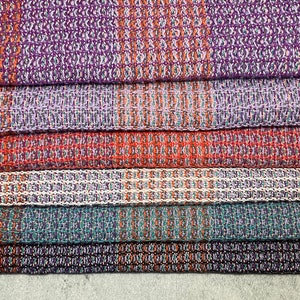 May include: A stack of six woven kitchen towels in various colors, including purple, orange, teal, and white. The towels have a textured pattern and are folded neatly.