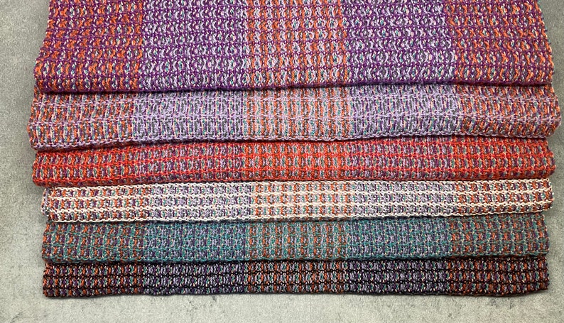 May include: A stack of six woven kitchen towels in various colors, including purple, orange, teal, and white. The towels have a textured pattern and are folded neatly.