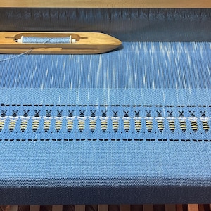 Puede incluir: Una tela tejida azul y blanca con un patrón de abejas en un telar. La tela se está tejiendo en un telar de madera con una lanzadera.