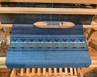 Puede incluir: Un telar de madera con una tela azul tejida que presenta un patrón repetitivo de abejas amarillas y negras. Una lanzadera de madera con una bobina de hilo azul descansa sobre los hilos de urdimbre sobre la sección tejida del proyecto.