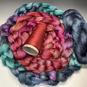 May include: A collection of colorful yarn in shades of teal, purple, pink, and red. A red spool of thread sits in the center of the yarn. The yarn is arranged in a circular pattern, creating a visually appealing display for crafting projects.