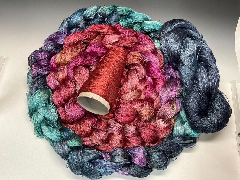 May include: A collection of colorful yarn in shades of teal, purple, pink, and red. A red spool of thread sits in the center of the yarn. The yarn is arranged in a circular pattern, creating a visually appealing display for crafting projects.