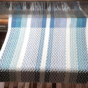 Puede incluir: Una pieza textil tejida en un telar con un patrón repetitivo de rayas verticales en tonos azul verdoso, azul marino y blanco. La tela muestra un tejido texturizado con bloques geométricos alternos y bandas de colores sólidos.