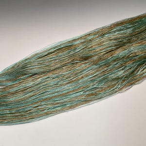Puede incluir: Primer plano de un ovillo de hilo con una mezcla de hilos azul claro y dorados. El hilo est&aacute; finamente hilado, con un ligero brillo, y parece suave. La combinaci&oacute;n de colores crea un efecto sutil y elegante, adecuado para proyectos de punto o ganchillo.