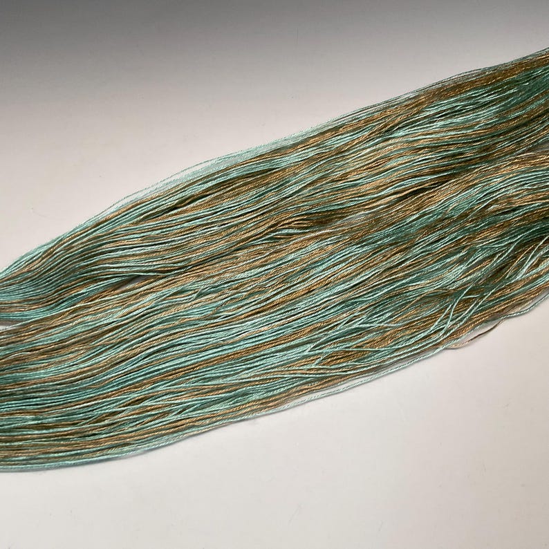 Puede incluir: Primer plano de un ovillo de hilo con una mezcla de hilos azul claro y dorados. El hilo est&aacute; finamente hilado, con un ligero brillo, y parece suave. La combinaci&oacute;n de colores crea un efecto sutil y elegante, adecuado para proyectos de punto o ganchillo.