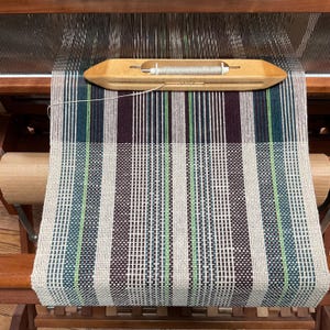 May include: A loom with a woven fabric in shades of white, brown, green, and blue. The fabric is being woven on a wooden loom with a shuttle and a bobbin.