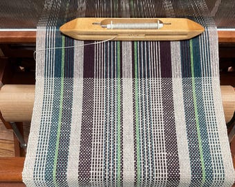 May include: A loom with a woven fabric in shades of white, brown, green, and blue. The fabric is being woven on a wooden loom with a shuttle and a bobbin.