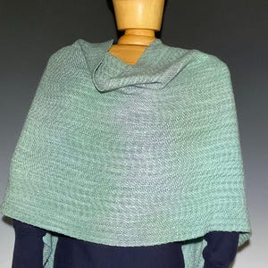 Pu&ograve; includere: Uno scialle verde chiaro tessuto drappeggiato su un manichino di legno. Lo scialle ha un aspetto strutturato e uno scollo a gorgiera. Il tessuto sembra essere un materiale morbido e leggero. Lo scialle &egrave; di colore uniforme e si drappeggia sulle spalle.
