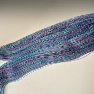 May include: A skein of hand-dyed yarn in shades of teal, purple, and white. The yarn appears to be a fine weight, with a subtle sheen. The colors are blended in a striped pattern, creating a visually appealing texture. Ideal for knitting or crochet projects.