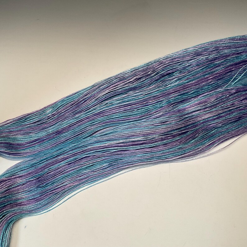 May include: A skein of hand-dyed yarn in shades of teal, purple, and white. The yarn appears to be a fine weight, with a subtle sheen. The colors are blended in a striped pattern, creating a visually appealing texture. Ideal for knitting or crochet projects.