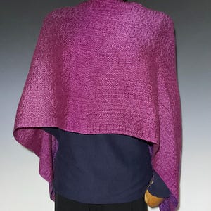 May include: A vibrant magenta shawl featuring a distinct herringbone weave pattern. The textile is draped over the shoulders, showcasing a textured surface with a consistent deep purple hue throughout the fabric, creating a structured appearance.