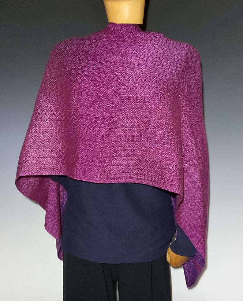 May include: A vibrant magenta shawl featuring a distinct herringbone weave pattern. The textile is draped over the shoulders, showcasing a textured surface with a consistent deep purple hue throughout the fabric, creating a structured appearance.
