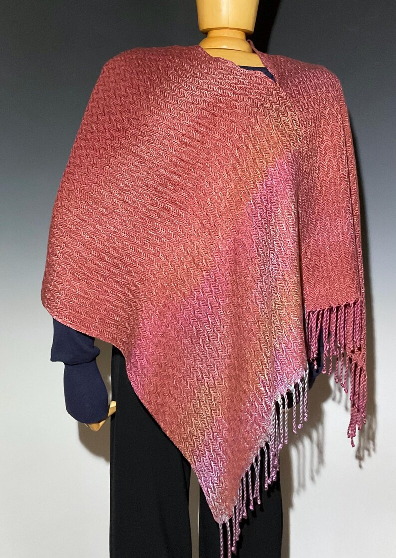May include: A woven shawl in shades of pink, peach, and lavender, displayed on a mannequin. The shawl has a textured, diagonal pattern and is finished with fringe along the edges. The garment is draped over a dark blue top and black pants.