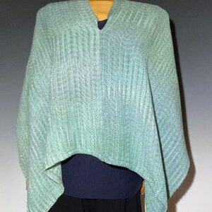 Pu&ograve; includere: Un poncho verde chiaro e blu con scollo a V. Il poncho in tessuto strutturato &egrave; esposto su un manichino di legno, sopra un top blu scuro e pantaloni neri. L'indumento ha un effetto sfumato.