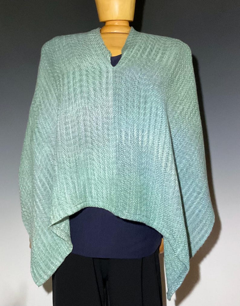 Pu&ograve; includere: Un poncho verde chiaro e blu con scollo a V. Il poncho in tessuto strutturato &egrave; esposto su un manichino di legno, sopra un top blu scuro e pantaloni neri. L'indumento ha un effetto sfumato.