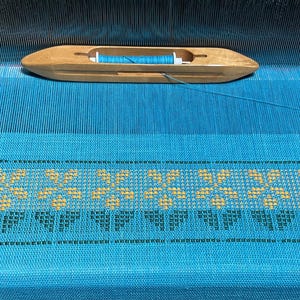 May include: A wooden shuttle with blue thread rests on a turquoise woven textile with a yellow floral pattern. The textile is on a wooden loom, with the warp threads visible. The image showcases the process of weaving.