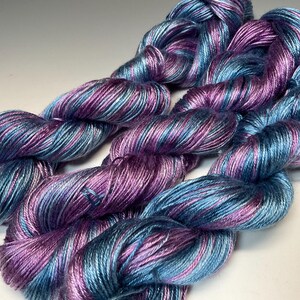 May include: Close-up of several skeins of yarn in shades of blue, purple, and pink. The yarn appears to be a fine weight, with a slight sheen. The colors are blended together in a variegated pattern, creating a visually appealing texture.