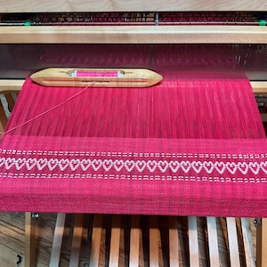 May include: A red woven fabric with a white heart pattern on a wooden loom. The fabric is being woven on a loom with a shuttle and a bobbin.