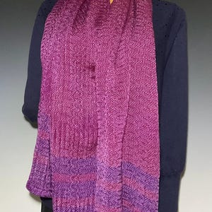 May include: A long, woven scarf featuring a vibrant magenta and purple herringbone pattern. The fabric displays a textured, zigzag weave throughout, with horizontal bands of deeper purple and magenta tones near the bottom edge of the garment.