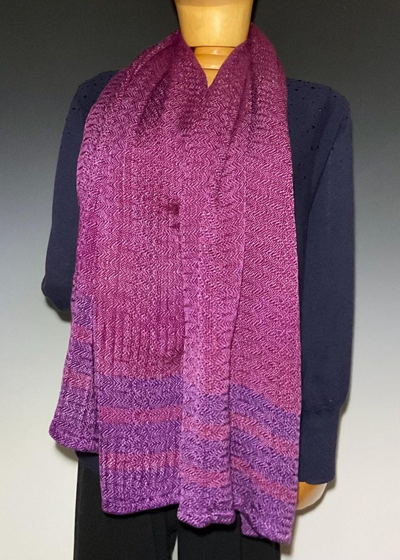 May include: A long, woven scarf featuring a vibrant magenta and purple herringbone pattern. The fabric displays a textured, zigzag weave throughout, with horizontal bands of deeper purple and magenta tones near the bottom edge of the garment.