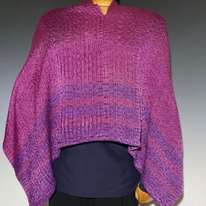 May include: A woven poncho featuring a vibrant magenta and purple herringbone pattern. The garment has a V-neckline and a draped silhouette, showcasing horizontal bands of darker purple stripes across the lower section of the fabric.
