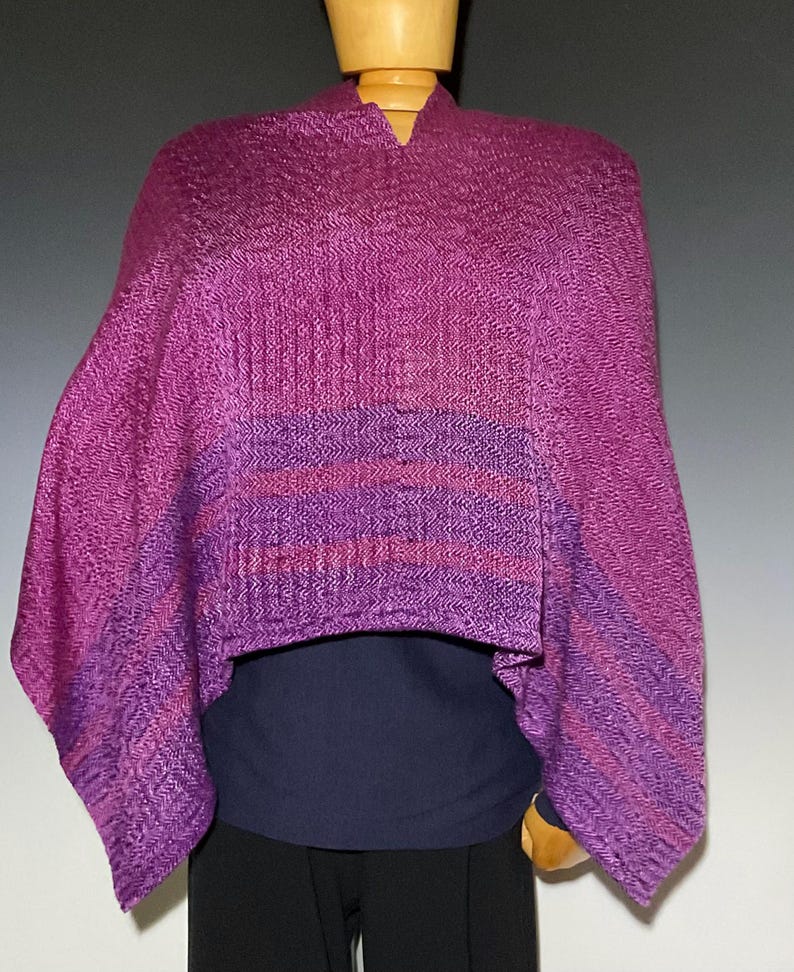 May include: A woven poncho featuring a vibrant magenta and purple herringbone pattern. The garment has a V-neckline and a draped silhouette, showcasing horizontal bands of darker purple stripes across the lower section of the fabric.