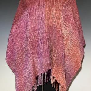 Op de afbeelding: Een geweven poncho met een kleurverloop van dieppaars en magenta naar warme oranje en koperkleurige tinten. Het kledingstuk heeft een gestructureerd visgraatpatroon over het geheel en is afgewerkt met franjes langs de onderzoom.