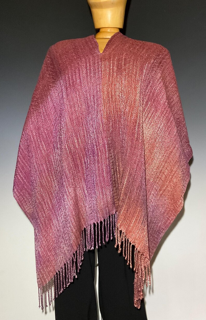 Op de afbeelding: Een geweven poncho met een kleurverloop van dieppaars en magenta naar warme oranje en koperkleurige tinten. Het kledingstuk heeft een gestructureerd visgraatpatroon over het geheel en is afgewerkt met franjes langs de onderzoom.
