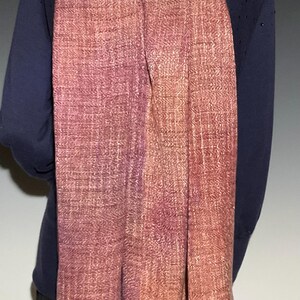 May include: A long, rectangular woven scarf featuring a gradient of colors transitioning from deep plum and purple tones on the left to warm, earthy shades of copper, rust, and muted orange on the right, with a visible textured weave pattern.