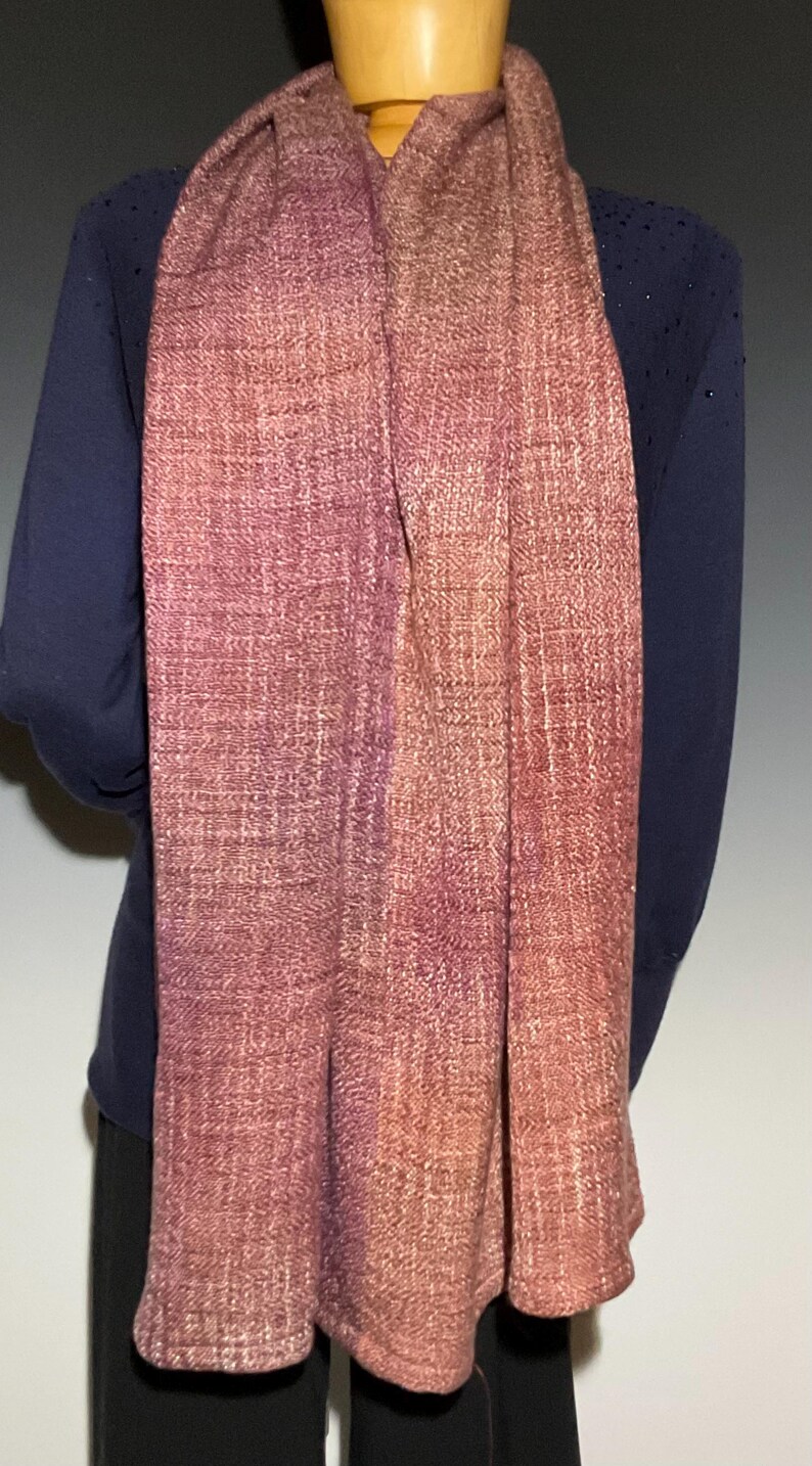 May include: A long, rectangular woven scarf featuring a gradient of colors transitioning from deep plum and purple tones on the left to warm, earthy shades of copper, rust, and muted orange on the right, with a visible textured weave pattern.