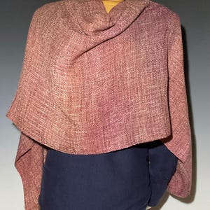 May include: A woven shawl in shades of dusty rose and mauve with a visible textured pattern. The fabric drapes over the shoulders, displaying a consistent weave throughout the material. The shawl is layered over a solid dark blue garment.