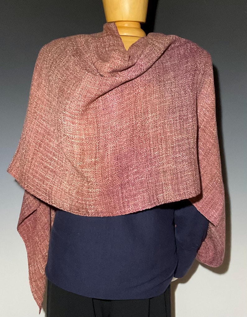 May include: A woven shawl in shades of dusty rose and mauve with a visible textured pattern. The fabric drapes over the shoulders, displaying a consistent weave throughout the material. The shawl is layered over a solid dark blue garment.