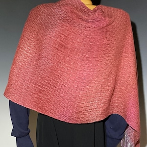 May include: A reddish-pink shawl with a woven pattern and fringed edges. The shawl is draped over a mannequin, showcasing its texture and design. The garment appears to be made of a soft, lightweight material.