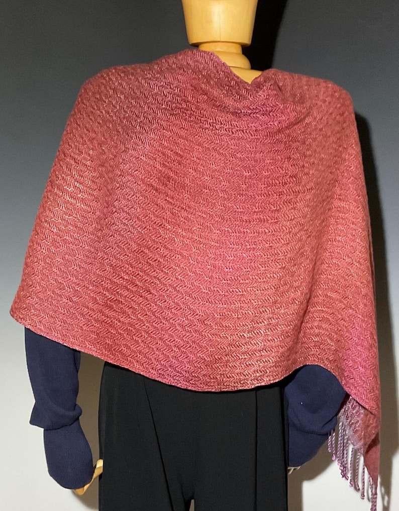 May include: A reddish-pink shawl with a woven pattern and fringed edges. The shawl is draped over a mannequin, showcasing its texture and design. The garment appears to be made of a soft, lightweight material.