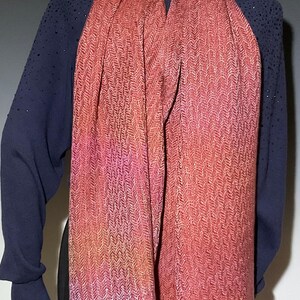 May include: A woven scarf in shades of red, pink, and orange with a herringbone pattern. The scarf has a fringed edge and is draped over a mannequin. The mannequin is wearing a navy blue long-sleeved top.