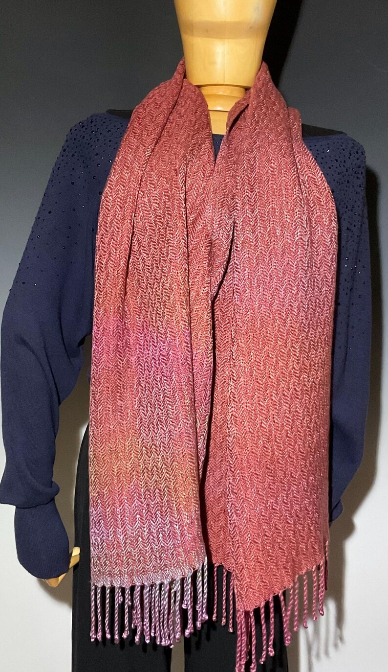 May include: A woven scarf in shades of red, pink, and orange with a herringbone pattern. The scarf has a fringed edge and is draped over a mannequin. The mannequin is wearing a navy blue long-sleeved top.
