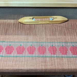 Puede incluir: Un telar con una tela tejida que presenta una fila de manzanas rojas con tallos y hojas verdes. La tela está tejida con hilo marrón y blanco. Una lanzadera de madera con un carrete de hilo marrón está en la parte superior del telar.