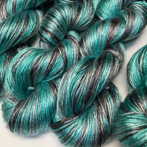 Op de afbeelding: Close-up van verschillende strengen garen in tinten teal, grijs en zwart. Het garen is strak gewikkeld, met een lichte glans. De kleurvariatie cre&euml;ert een gemarmerd effect, geschikt voor brei- of haakprojecten.