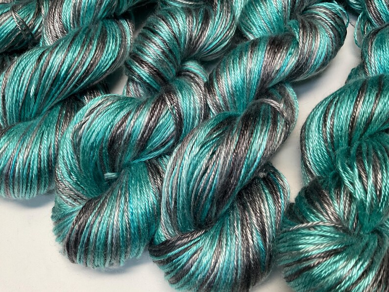 Op de afbeelding: Close-up van verschillende strengen garen in tinten teal, grijs en zwart. Het garen is strak gewikkeld, met een lichte glans. De kleurvariatie cre&euml;ert een gemarmerd effect, geschikt voor brei- of haakprojecten.