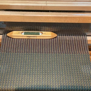 May include: A close-up of a loom with a green and brown striped fabric being woven. A wooden shuttle with a green thread is visible in the foreground.