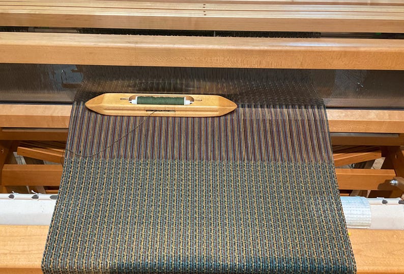 May include: A close-up of a loom with a green and brown striped fabric being woven. A wooden shuttle with a green thread is visible in the foreground.
