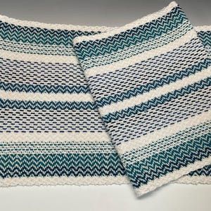 May include: A rectangular woven table runner featuring a repeating pattern of horizontal stripes in cream, teal, and dark blue. The design incorporates alternating sections of solid bands and intricate chevron-style zig-zag weave textures.