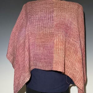 May include: A woven poncho featuring a variegated color palette of mauve, dusty rose, and soft orange tones. The fabric displays a textured, cross-hatched weave pattern throughout. The garment is draped over a mannequin, layered above a dark blue top.
