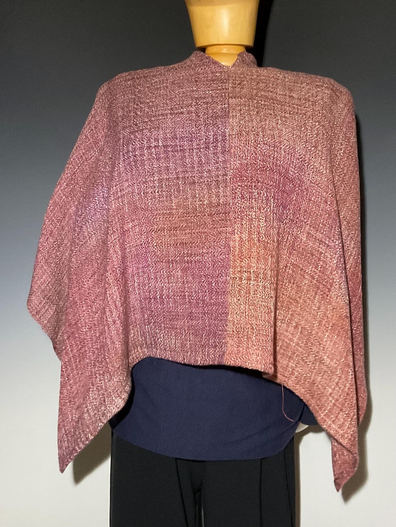 May include: A woven poncho featuring a variegated color palette of mauve, dusty rose, and soft orange tones. The fabric displays a textured, cross-hatched weave pattern throughout. The garment is draped over a mannequin, layered above a dark blue top.
