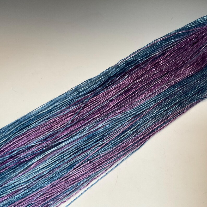 May include: Close-up of a bundle of yarn with a gradient of colors. The yarn transitions from a light blue to a deep purple hue. The yarn is tightly spun and appears soft, ideal for knitting or crocheting projects.
