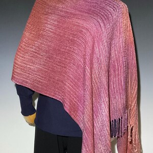 Op de afbeelding: Een geweven sjaal met een kleurverloop in roze, magenta en koperkleurige tinten met een textuur van ribbels. Het kledingstuk valt over de schouders en is voorzien van franjes aan de onderkant, gedragen over een donkerblauw shirt met lange mouwen.