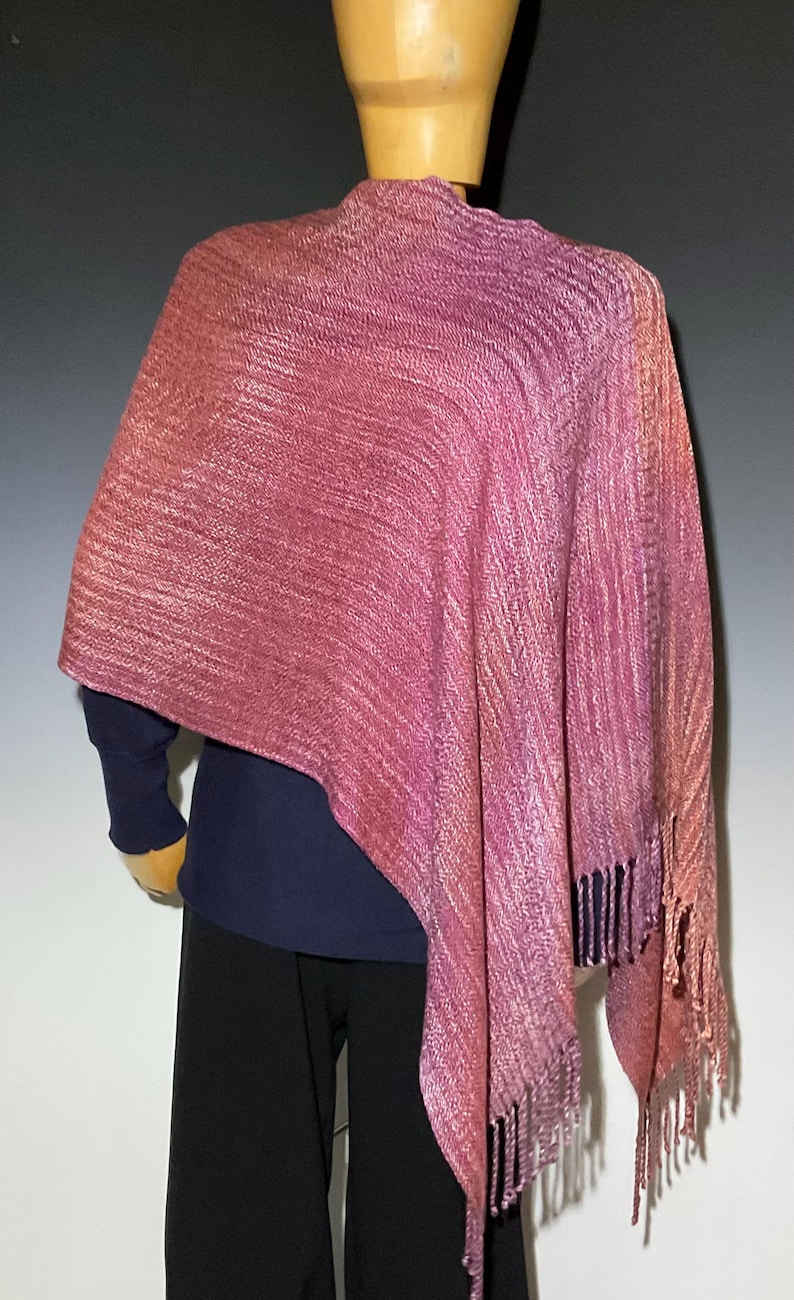 Op de afbeelding: Een geweven sjaal met een kleurverloop in roze, magenta en koperkleurige tinten met een textuur van ribbels. Het kledingstuk valt over de schouders en is voorzien van franjes aan de onderkant, gedragen over een donkerblauw shirt met lange mouwen.