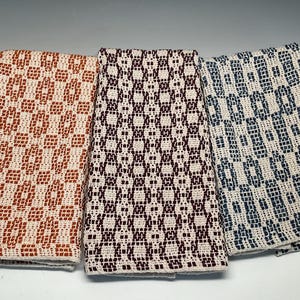 May include: Three rectangular woven kitchen towels featuring a geometric pattern. The left towel is orange and cream, the centre towel is dark maroon and cream, and the right towel is navy blue and cream. Each towel displays a repeating textured weave.