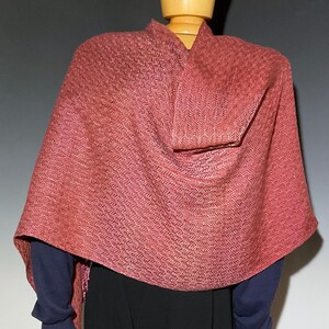 May include: A woven shawl in shades of red and pink, draped over a mannequin. The shawl features a textured, herringbone pattern. The mannequin is wearing a dark blue long-sleeved shirt and a black skirt.